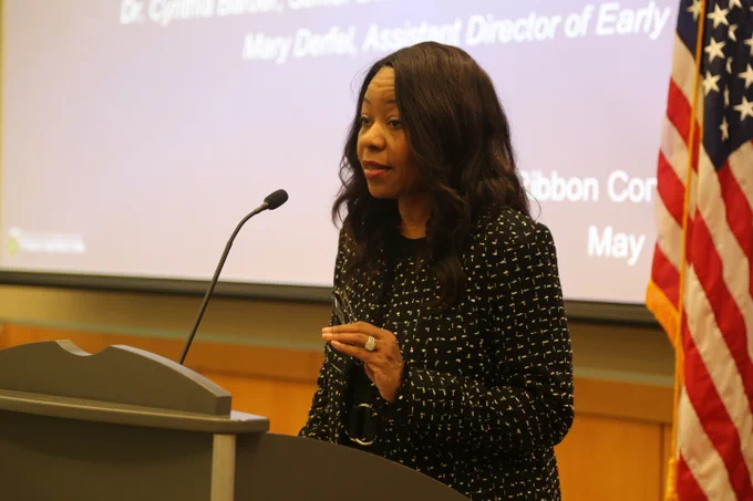 Woman in a patterned black blazer speaking at a podium with a microphone in a formal setting.