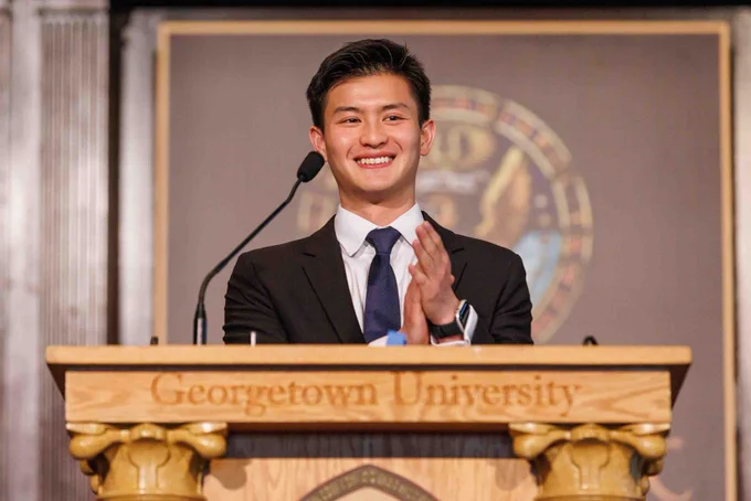 An individual standing at a podium with "Georgetown University" inscribed on it, smiling and clapping.