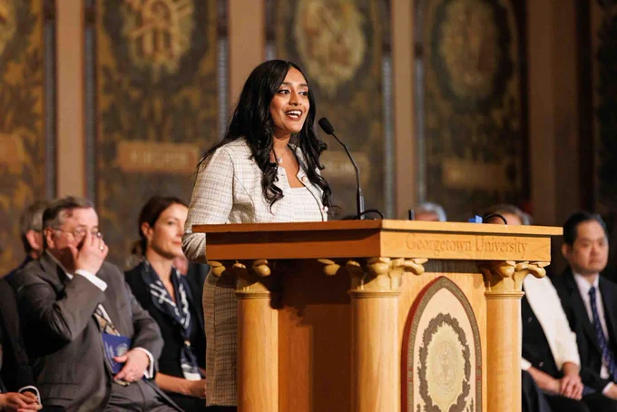 A person speaking at a podium at Georgetown University. The audience includes several individuals seated, listening attentively. The background features ornate architectural details.