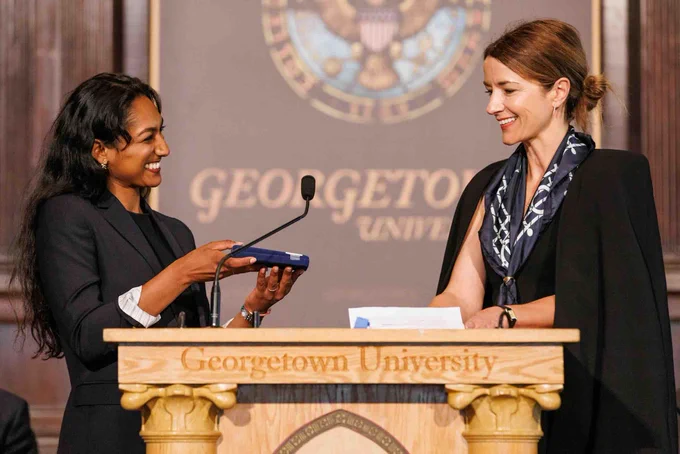 Two people stand at a podium with a Georgetown University emblem in the background. One person is holding an award and smiling at the other.