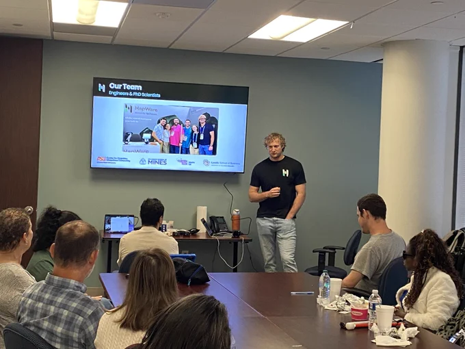 A person is giving a presentation to a group in a conference room. A screen displays information about engineers and scientists, with logos of Hines, HOK, and several universities. Attendees are seated around a table with laptops and beverages.
