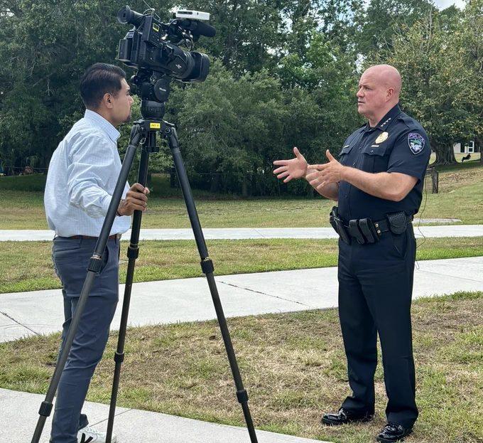 A news reporter interviews a bald police officer outdoors. The reporter stands next to a camera on a tripod, wearing a light blue shirt and dark pants. The officer, dressed in a dark uniform with a badge and equipment belt, gestures with his hands while speaking. They are situated on a grassy area with a sidewalk and trees in the background.