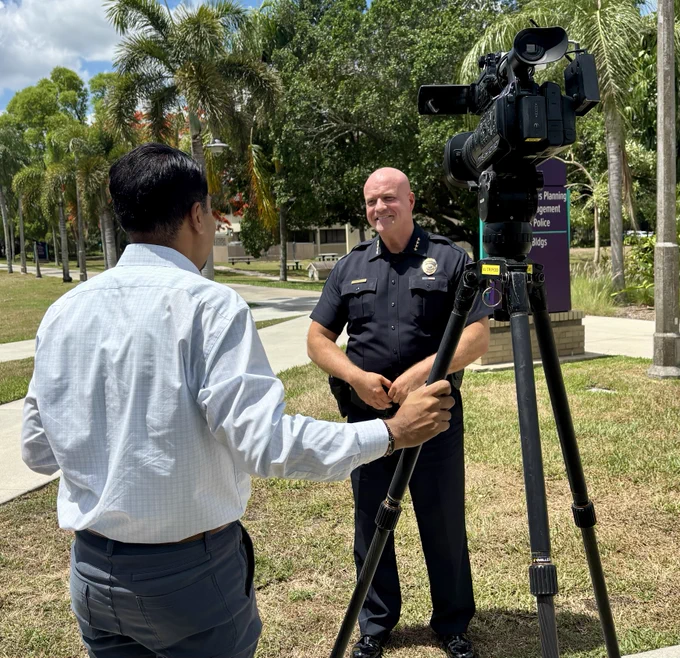 A man in a light blue shirt holds a video camera on a tripod, filming a bald man in a police uniform outside. The setting features a walkway and palm trees under a partly cloudy sky.