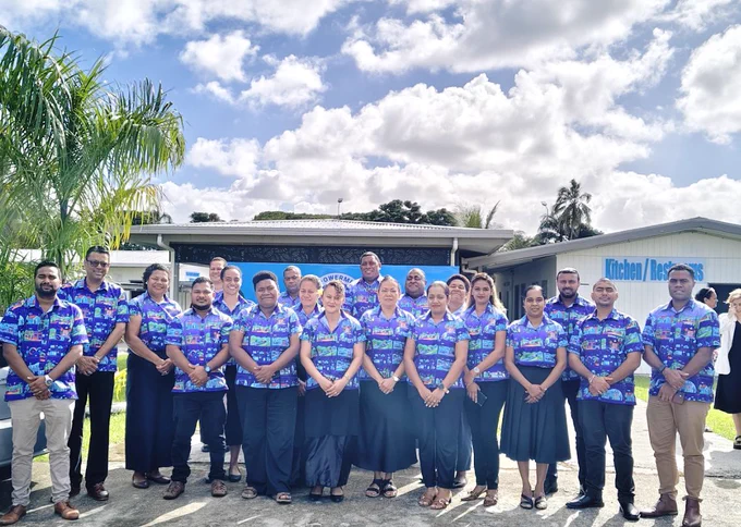 Group photo of participants wearing matching blue patterned shirts standing outdoors in front of the FLIPS training building under a partly cloudy sky.