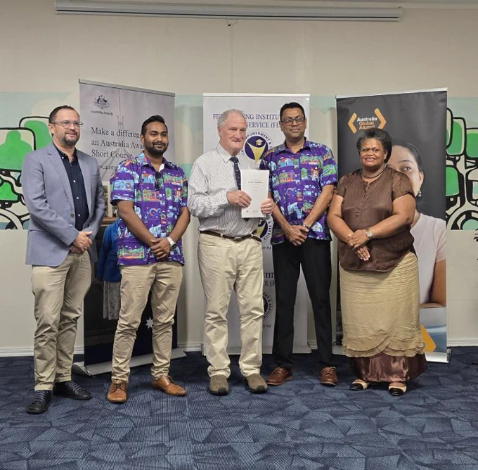 Participants and course staff stand indoors in front of banners, with one person in the centre holding a certificate of completion, dressed in business casual and traditional attire.