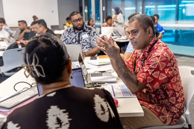 A group of participants engaged in a collaborative discussion around a table with laptops and documents in a modern office setting.
