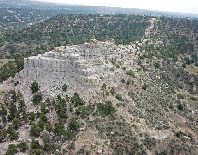 Aerial view of Castle Rock, a prominent rock formation surrounded by greenery and sparse vegetation. The horizon shows a vast landscape extending under an overcast sky.