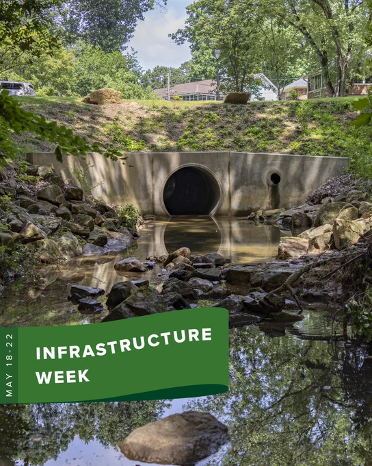A culvert with water flowing through it is surrounded by rocks and lush greenery. A house is visible in the background. A green banner reads "Infrastructure Week, May 18-22."