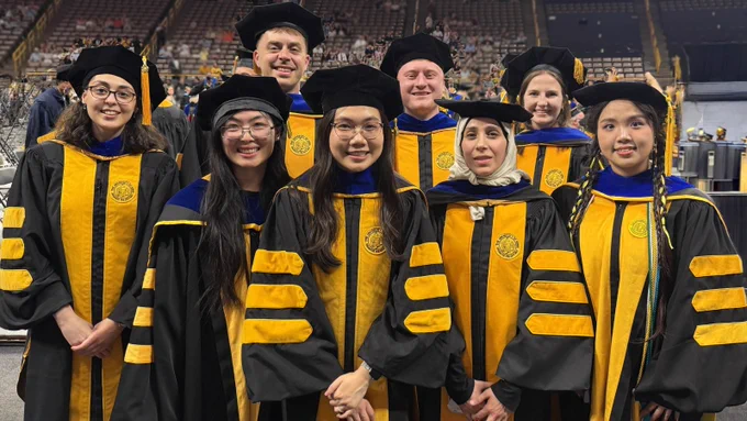 Eight University of Iowa College of Pharmacy graduates in black doctoral regalia with gold accents pose together on the commencement floor inside Carver-Hawkeye Arena. The graduates smile toward the camera while standing among other attendees before the ceremony.