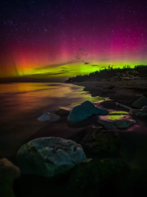 Vibrant aurora borealis above a serene beach with colorful green and pink lights reflecting on the water, creating a mystical, tranquil atmosphere.