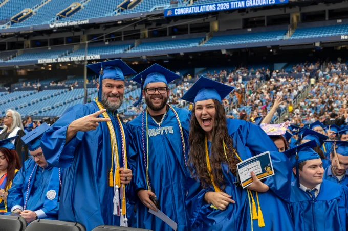 A group of graduates wearing blue caps and gowns are celebrating at a graduation ceremony, posing with smiles and laughing. One individual is making a peach sign with their hand. They are standing in a stadium.