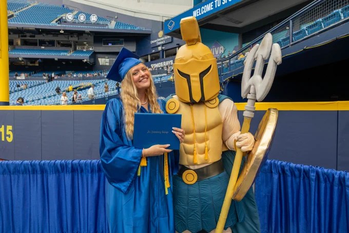 A person in a blue graduation gown and cap holds a diploma, standing next to SPC mascot Titus. They are in a sports stadium, with Tampa Bay Rays logos visible in the background.