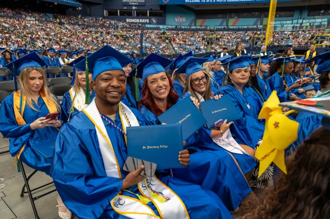 A group of graduates in blue caps and gowns are seated in a large stadium filled with people. They are holding diplomas, smiling, and celebrating their achievement.