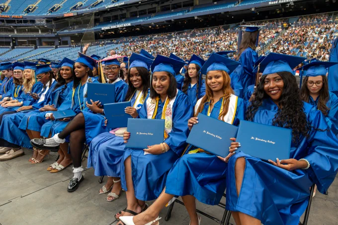 Graduates seated in a stadium wearing blue cap and gowns, each holding SPC diplomas. The background shows an audience in the stands.