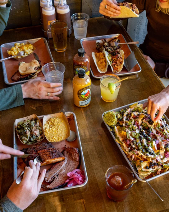 Four people share a meal at a wooden table with barbecue meats, tacos, nachos, corn, drinks, and sauces. Hands are reaching for food, creating a casual, lively dining scene.
