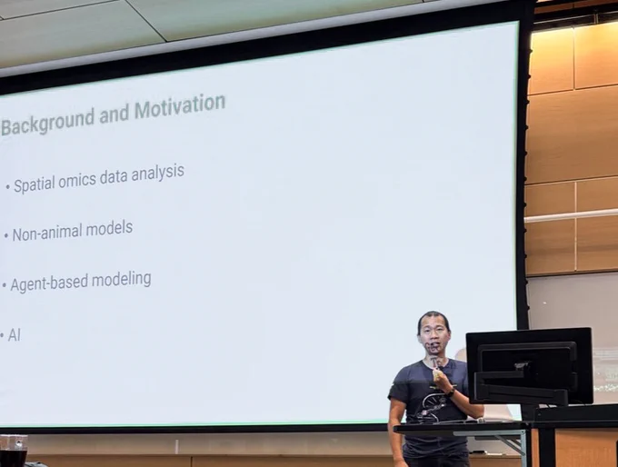 A presenter stands at a podium in a lecture hall beside a large projection screen displaying a slide titled “Background and Motivation.” The slide lists bullet points including spatial omics data analysis, non-animal models, agent-based modeling, and AI.