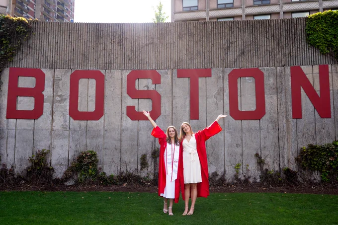 Photo of Anastasiia Semenova (left) and Anete Adler (right) in their graduation gowns in front of the Boston University sign at Nickerson Field