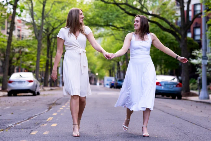 Photo of Anete Adler (left) and Anastasiia Semenova (right) wearing white dresses while walking down the street holding hands.