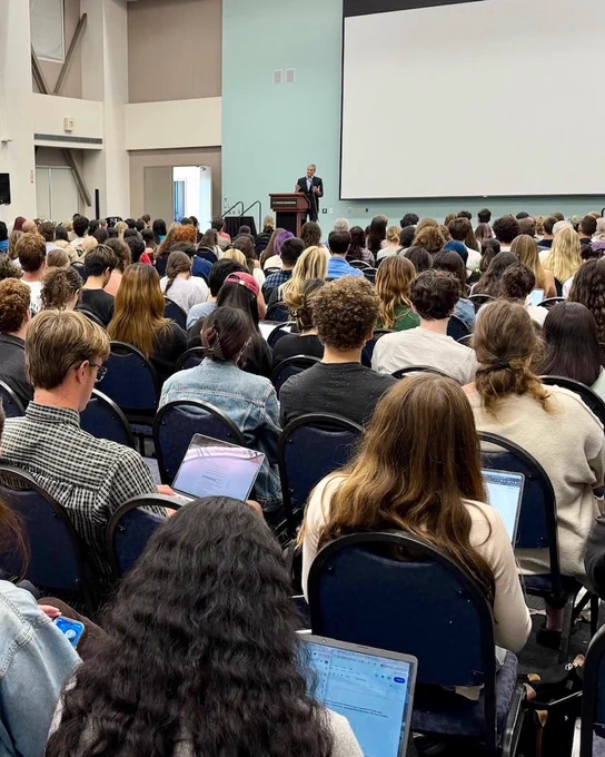 A large audience is seated in rows in a lecture hall, listening to Wade Crowfoot present on stage next to a large projector screen.