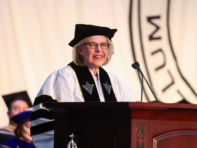 A person in academic regalia speaks at a podium during a commencement ceremony. The backdrop includes a large banner.