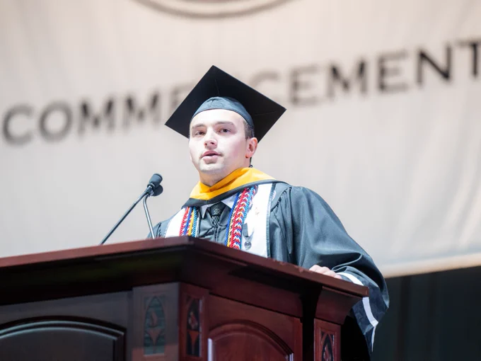 A person in graduation attire stands at a podium speaking into a microphone. A banner behind them displays the partial word "Commencement."