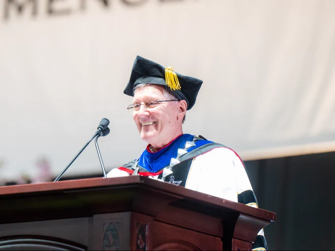 An individual wearing academic regalia, including a cap with a tassel, stands at a podium with a microphone, smiling. The setting is a graduation.