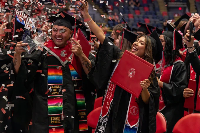 Graduates in black gowns and red stoles celebrate as red and white confetti falls. Several hold red diploma folders, including one with visible text reading “Class of 2026,” while others raise their hands.