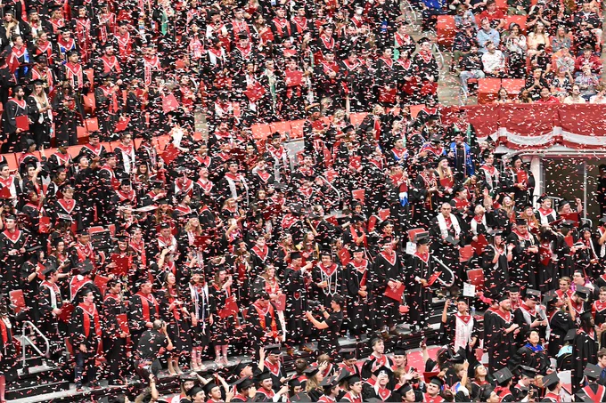 Beasley Coliseum filled with graduates wearing black gowns and red stoles, many holding red diplomas. Red and white confetti falls across the scene while spectators watch from the stands.