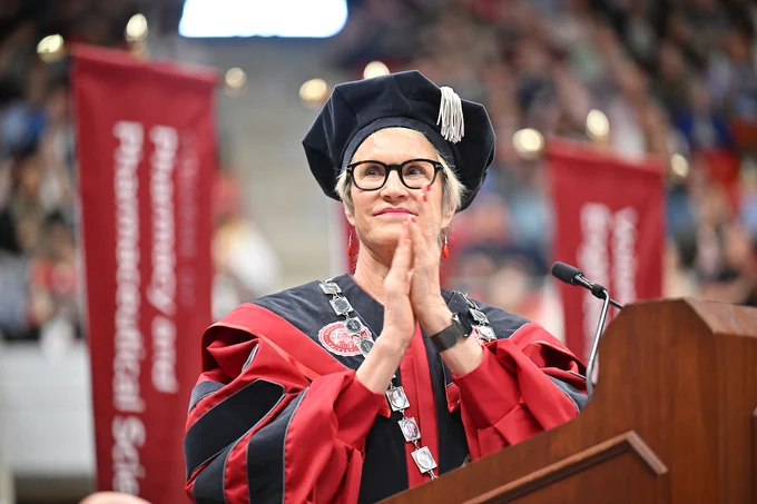 President Cantwell stands at a podium in red and black academic regalia and a doctoral cap. A microphone and red banners appear nearby, with a blurred audience in a large indoor venue behind them.