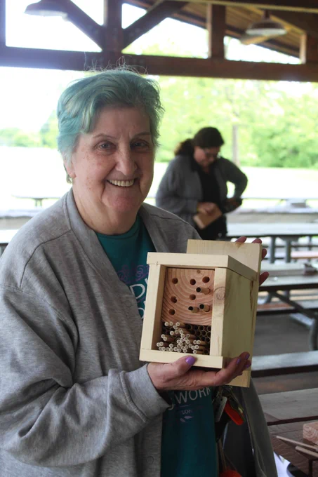 A woman holds up a completed bee hotel - a wood box with one side open that has blocks of woods with drilled holes and straws.