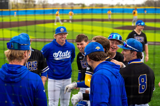 South Dakota State baseball players gather in a circle on the field, smiling and laughing during pregame warmups. Players wear blue and black Jackrabbits gear with sunglasses and caps, while a baseball diamond is visible in the background.