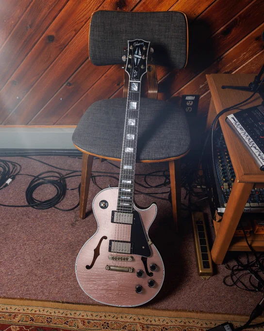 A Gibson Les Paul Custom with F-holes guitar with a sparkle pink finish rests against a gray cushioned chair. The background features a wooden paneled wall and music equipment, including cables and a mixing console.