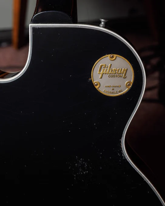 The image shows a close-up of a black Gibson guitar body with a circular gold emblem. The emblem reads "Gibson Custom" and "Hand Crafted in Nashville, TN.