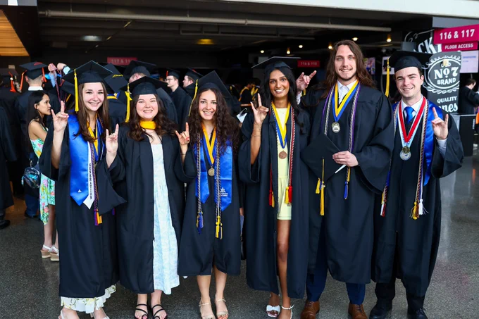 Six graduates stand together and smile for a photo, holding their hands in a Roo Up gesture.