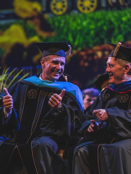 Two people in graduation gowns and caps are sitting side by side, smiling and giving thumbs up. They are on a stage with a blurred background, which includes greenery. 