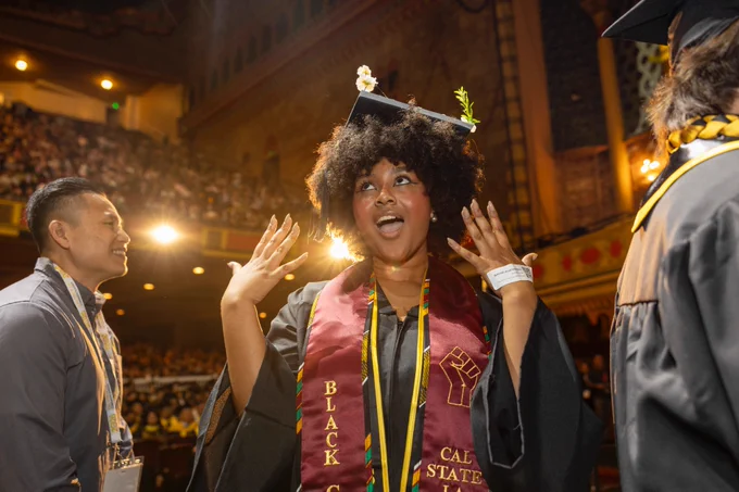 A graduate wearing a cap and gown with a "Cal State" stole celebrates joyfully at a ceremony. The background shows an audience in a theater-like venue.
