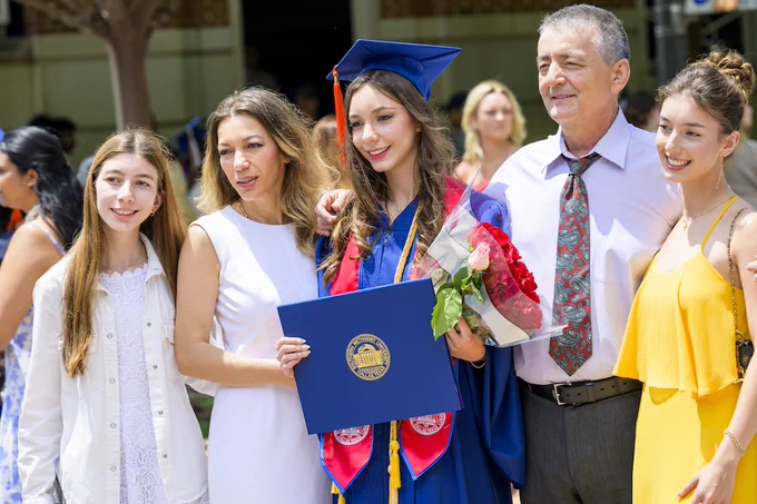 An SMU Lyle graduate in cap and gown holds a blue diploma folder and a bouquet of red roses, standing with four people. They are outdoors with a crowd in the background.