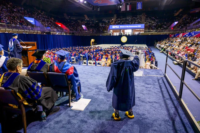 Buzz the robot stands in a cap and gown on stage during SMU Lyle's graduation ceremony, facing a large audience in a stadium. 