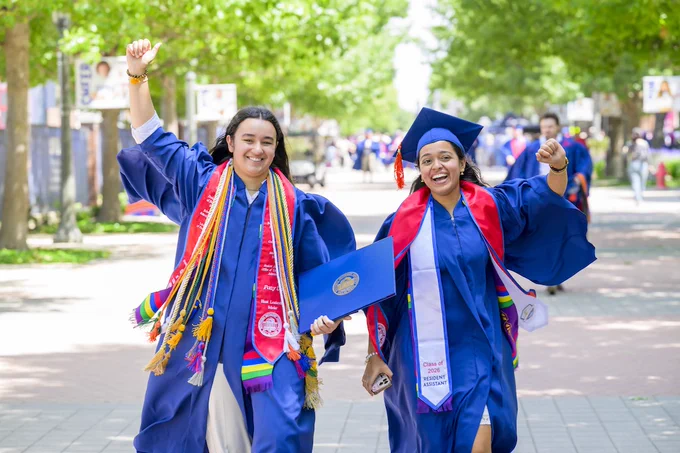 Two SMU Lyle students in graduation attire celebrate while walking on a tree-lined path. They wear blue gowns with colorful sashes and hold diplomas. The scene is festive and joyful, indicating a graduation event.