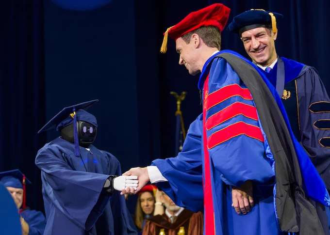 A graduate shakes hands with Buzz the robot while the SMU Lyle dean looks on and smiles