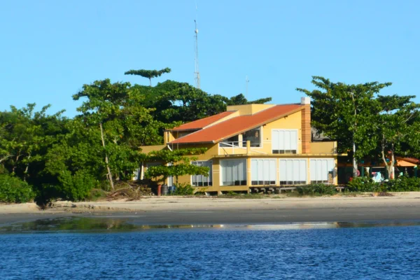 Foto colorida mostra uma casa de veraneio de dois pavimentos construída ilegalmente na Ilha das Peças (PR); com paredes amarelas e amplas janelas de vidro na fachada voltada para a praia, a casa está posicionada diretamente na faixa de areia da praia, de frente para a água mansa do mar, que ocupa o primeiro plano da foto sob um céu azul e limpo. Crédito: MPF.