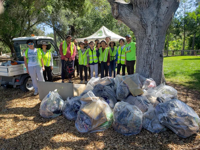 A group of people wearing high-visibility vests stand together in a park after a cleanup event. In front of them are numerous bags filled with collected trash. A large tree and a grassy area are in the background.