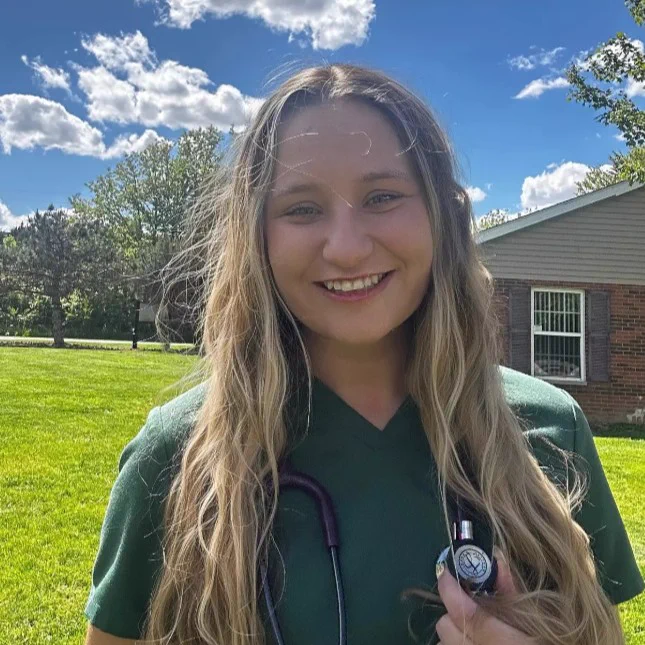 Hayley Bradbury smiling outdoors on a sunny day. They are wearing a green top and have a stethoscope around their neck. The background features a grassy area, trees, a building, and a sky filled with fluffy clouds.