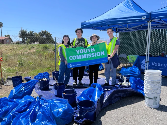 Four people are standing under a blue canopy tent, holding a "Youth Commission" sign. They are surrounded by blue garbage bags and buckets on a tarp. The setting is outdoors on a sunny day.