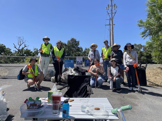 A group of people in high-visibility vests and hats are posing with litter-picking tools. They stand and sit on a pathway near a collection of trash bags. Behind them are trees and a clear blue sky. A table in the foreground holds snacks, drinks, and a water bottle.