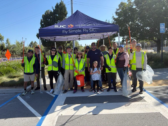 A group of people wearing reflective vests stand together in front of a tent with the "SJC" logo and the words "Fly Simple. Fly SJC." They are holding trash grabbers and buckets, suggesting participation in a community cleanup event. Trees and a parking sign are visible in the background.