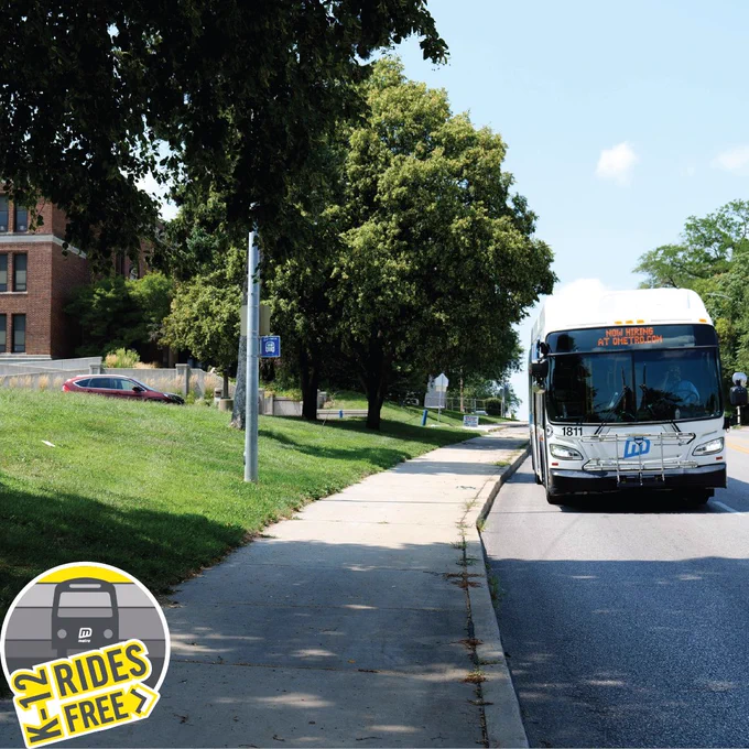 Photo of a Metro Transit white and blue bus stopped at a bus stop next to a school. Logo in bottom left reads, "K-12 rides free".