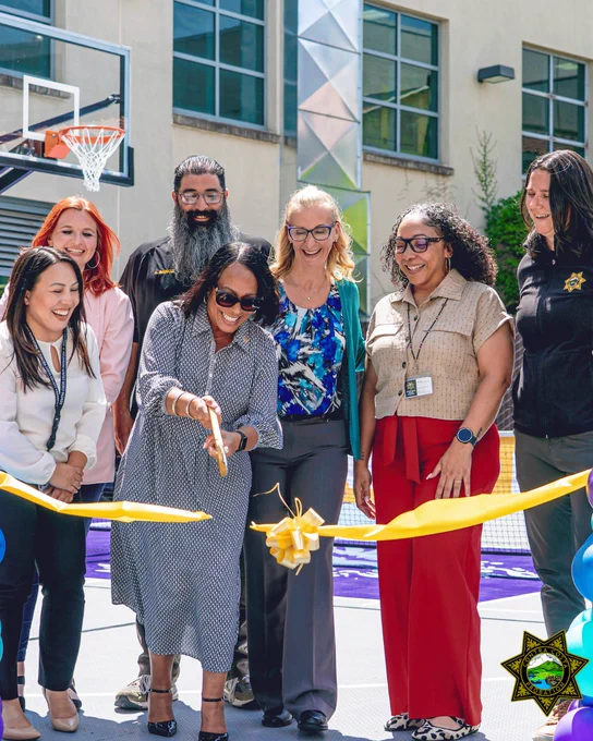 A group of eight people smiles as a woman cuts a yellow ribbon at a ceremony on a basketball court. Colorful balloons and a sunny day add cheer.