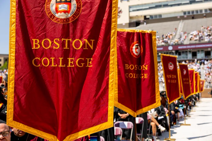 Red banners with "Boston College" text are displayed in a row, featuring the university's emblem. The scene appears to be a graduation ceremony in a large stadium with many attendees seated.