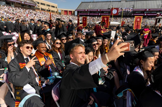 A group of graduates in caps and gowns sit in outdoor stadium seating during a commencement ceremony at a university. One individual in the foreground is taking a selfie. The stands are filled with spectators.